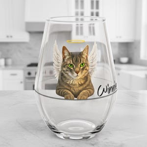 May include: A clear stemless wine glass decorated with a tabby cat design. The cat has angel wings, a halo, and green eyes. The name "Winnie" is written in cursive. The glass is on a white surface.