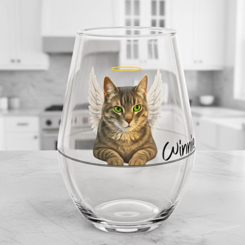 May include: A clear stemless wine glass decorated with a tabby cat design. The cat has angel wings, a halo, and green eyes. The name "Winnie" is written in cursive. The glass is on a white surface.