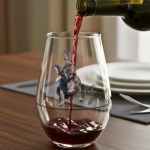 May include: A clear wine glass with a French Bulldog illustration, being filled with red wine from a bottle. The dog is grey and white, wearing a blue jacket. The glass has a black line and script text.