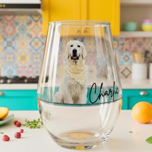 May include: A clear, stemless wine glass with the name "Charlie" in black script. Inside the glass is a Golden Retriever wearing a yellow bandana with white daisy accents. The dog is sitting in a turquoise-coloured liquid.