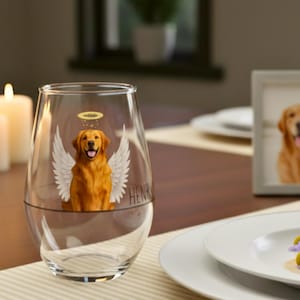 May include: A clear stemless wine glass featuring a golden retriever angel illustration with a halo and wings. A framed photo of a golden retriever and lit candles are on the table. The word "HENRY" is on the glass.