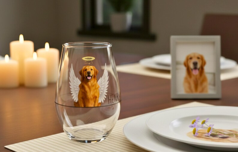 May include: A clear stemless wine glass featuring a golden retriever angel illustration with a halo and wings. A framed photo of a golden retriever and lit candles are on the table. The word "HENRY" is on the glass.