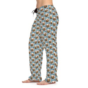 Pode incluir: Calças de pijama azul claro com um padrão repetido de rostos de gatos de desenho animado em torradas. Os gatos têm rostos pretos, olhos amarelos e estão em torradas bege. As calças têm um cordão preto e um ajuste folgado.