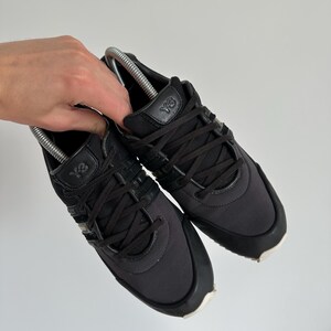 May include: A pair of black Y-3 sneakers with black laces and a white sole. The sneakers have the Y-3 logo on the tongue and side. The shoes are held by a hand.