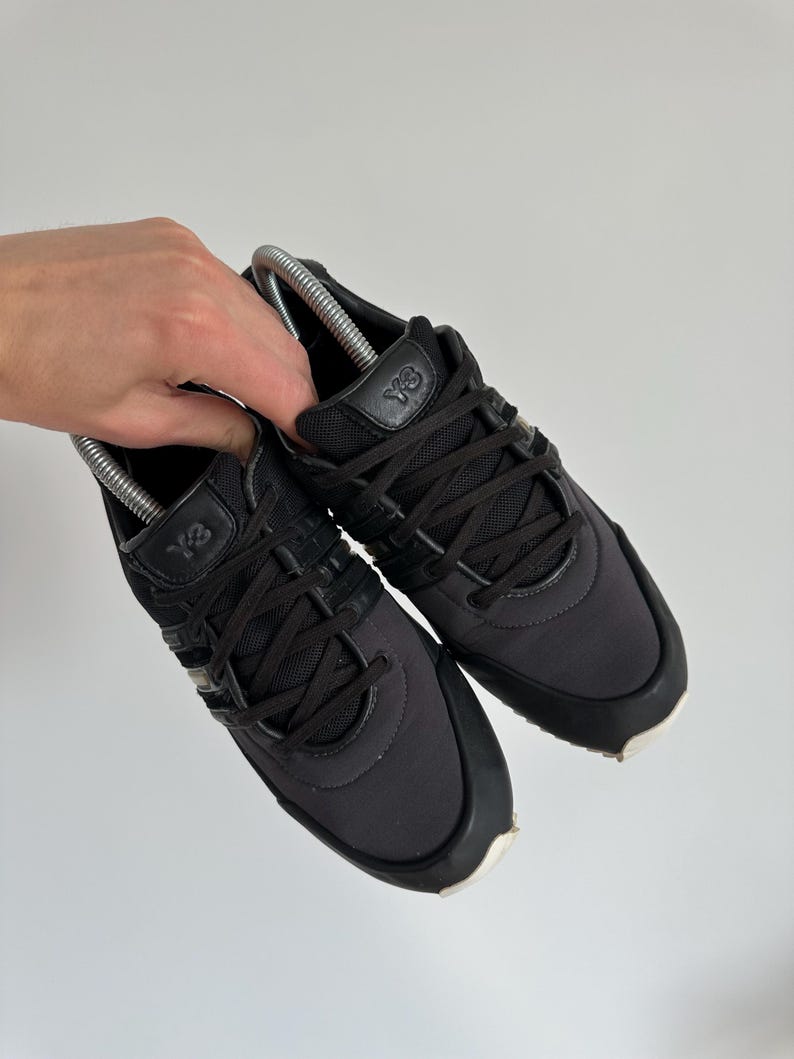 May include: A pair of black Y-3 sneakers with black laces and a white sole. The sneakers have the Y-3 logo on the tongue and side. The shoes are held by a hand.
