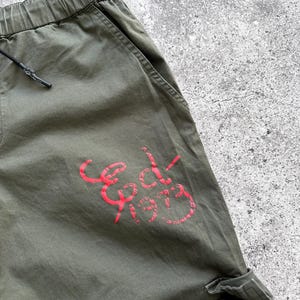 May include: Olive green cargo pants with an elastic waistband and drawstring. The pants feature a red graphic that reads "Ed-1973" on the leg. The pants have pockets and a relaxed fit.