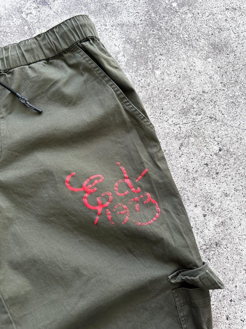 May include: Olive green cargo pants with an elastic waistband and drawstring. The pants feature a red graphic that reads "Ed-1973" on the leg. The pants have pockets and a relaxed fit.