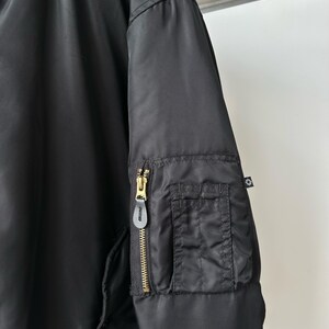 May include: A black bomber jacket featuring a smooth nylon-like fabric, a ribbed collar, and a utility pocket on the sleeve. The pocket includes a gold-toned metal zipper with a black leather pull tab and a small black fabric tag with a white logo.