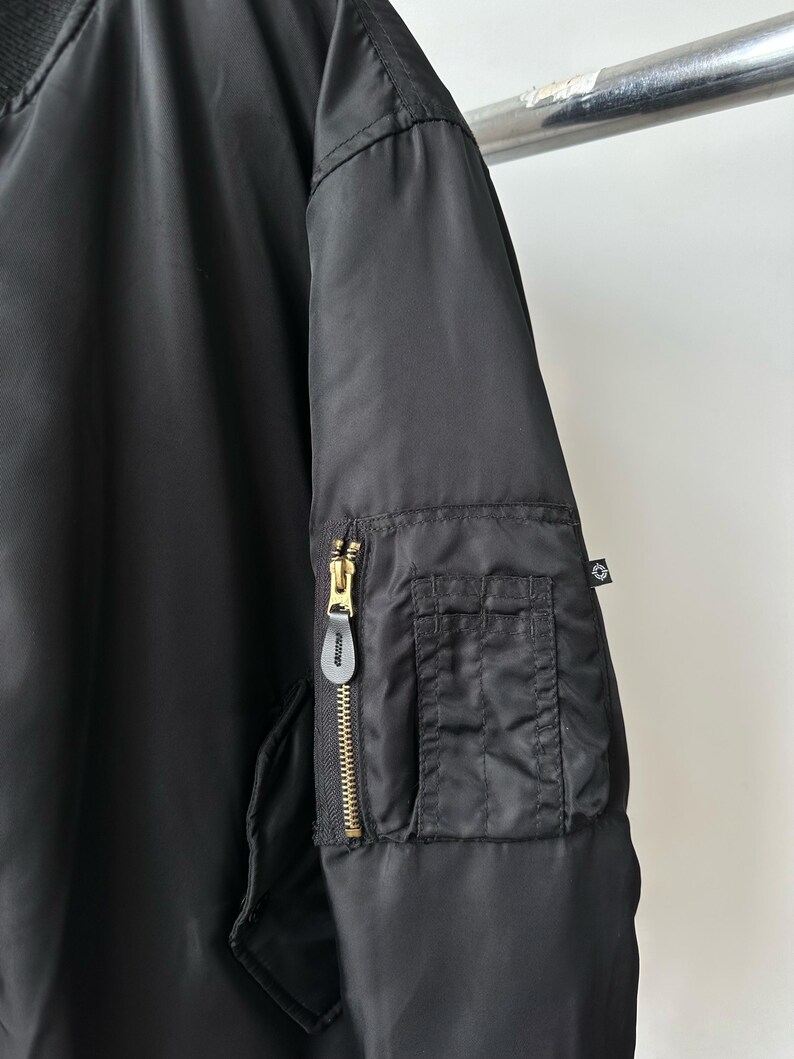 May include: A black bomber jacket featuring a smooth nylon-like fabric, a ribbed collar, and a utility pocket on the sleeve. The pocket includes a gold-toned metal zipper with a black leather pull tab and a small black fabric tag with a white logo.