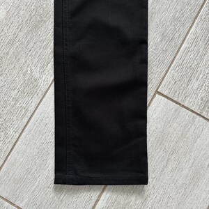 May include: A close-up view of a black pant leg featuring a straight-cut design. The fabric is a solid black color with visible vertical stitching along the side seam and a horizontal hem stitch at the bottom edge of the garment leg.