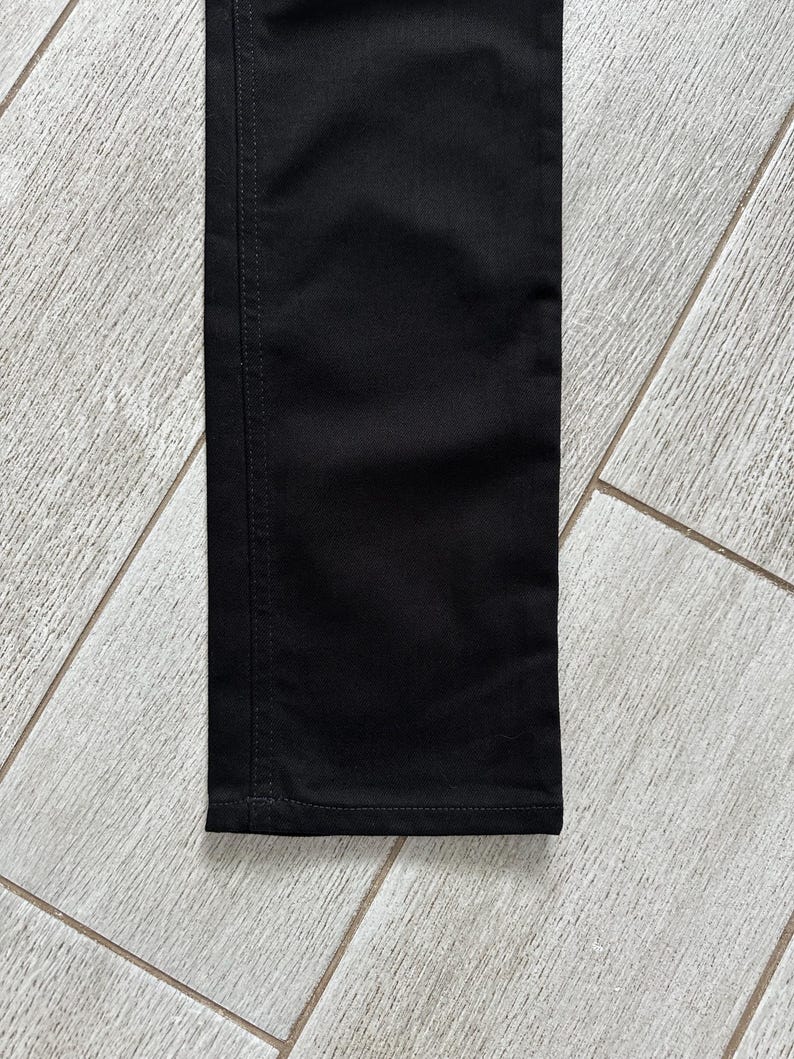 May include: A close-up view of a black pant leg featuring a straight-cut design. The fabric is a solid black color with visible vertical stitching along the side seam and a horizontal hem stitch at the bottom edge of the garment leg.
