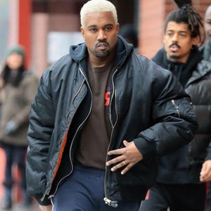 May include: A black bomber jacket with a silver zipper and orange lining, worn over a dark brown hooded sweatshirt. The outfit includes navy blue sweatpants with white text on the leg. The person has short, bleached blonde hair and a goatee.