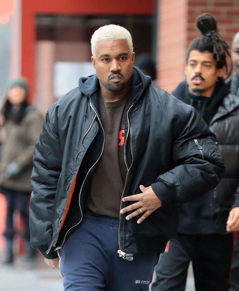 May include: A black bomber jacket with a silver zipper and orange lining, worn over a dark brown hooded sweatshirt. The outfit includes navy blue sweatpants with white text on the leg. The person has short, bleached blonde hair and a goatee.