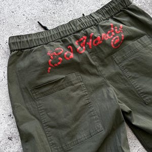 May include: Olive green cargo pants with a drawstring waist and two back pockets. The red text "Ed Hardy" is printed across the back waistband. The pants are made of a durable fabric.
