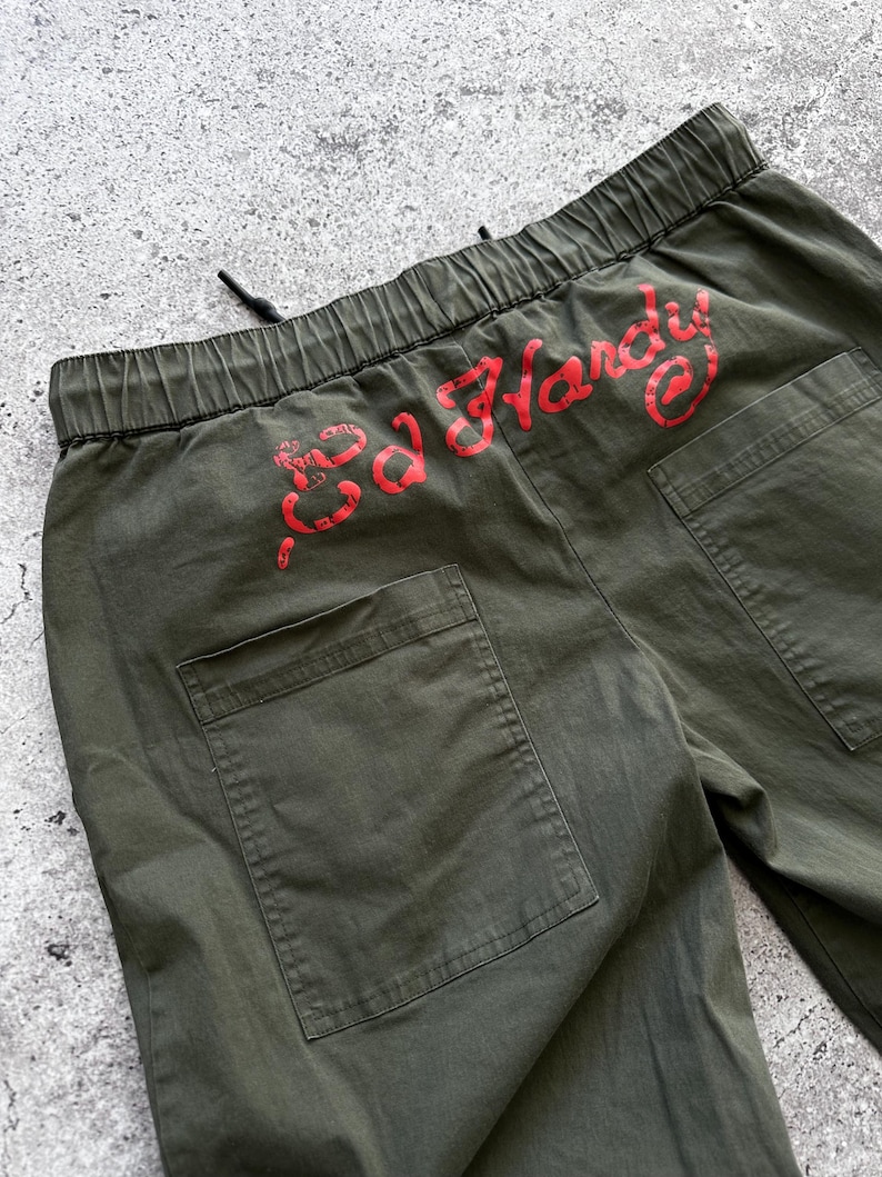 May include: Olive green cargo pants with a drawstring waist and two back pockets. The red text "Ed Hardy" is printed across the back waistband. The pants are made of a durable fabric.