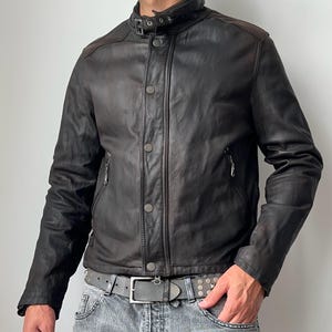 May include: A dark brown leather jacket featuring a stand-up collar with a buckle strap, a front button placket, and two zippered side pockets. The jacket has long sleeves and a smooth finish, paired with a grey belt and light grey denim jeans.