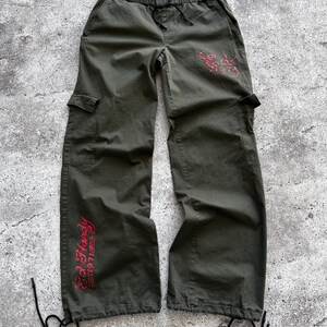 May include: Olive green cargo pants with a drawstring waist and multiple pockets. The pants feature red text that reads "Ed Hardy 1971" on the leg and a design on the back pocket. The pants have drawstrings at the ankles.