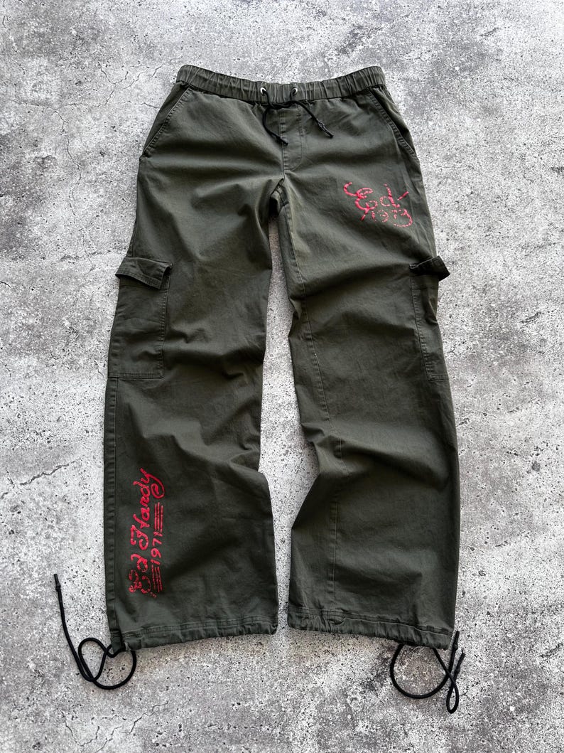 May include: Olive green cargo pants with a drawstring waist and multiple pockets. The pants feature red text that reads "Ed Hardy 1971" on the leg and a design on the back pocket. The pants have drawstrings at the ankles.