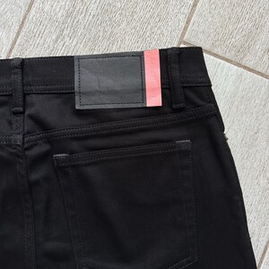 May include: Black denim jeans featuring a rear patch pocket and a black leather waistband label with a pink accent strip. The label displays the text Acne Studios in a subtle embossed font. The garment includes belt loops and tonal stitching.