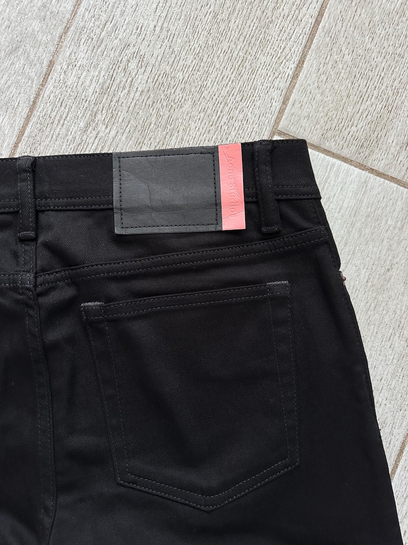 May include: Black denim jeans featuring a rear patch pocket and a black leather waistband label with a pink accent strip. The label displays the text Acne Studios in a subtle embossed font. The garment includes belt loops and tonal stitching.