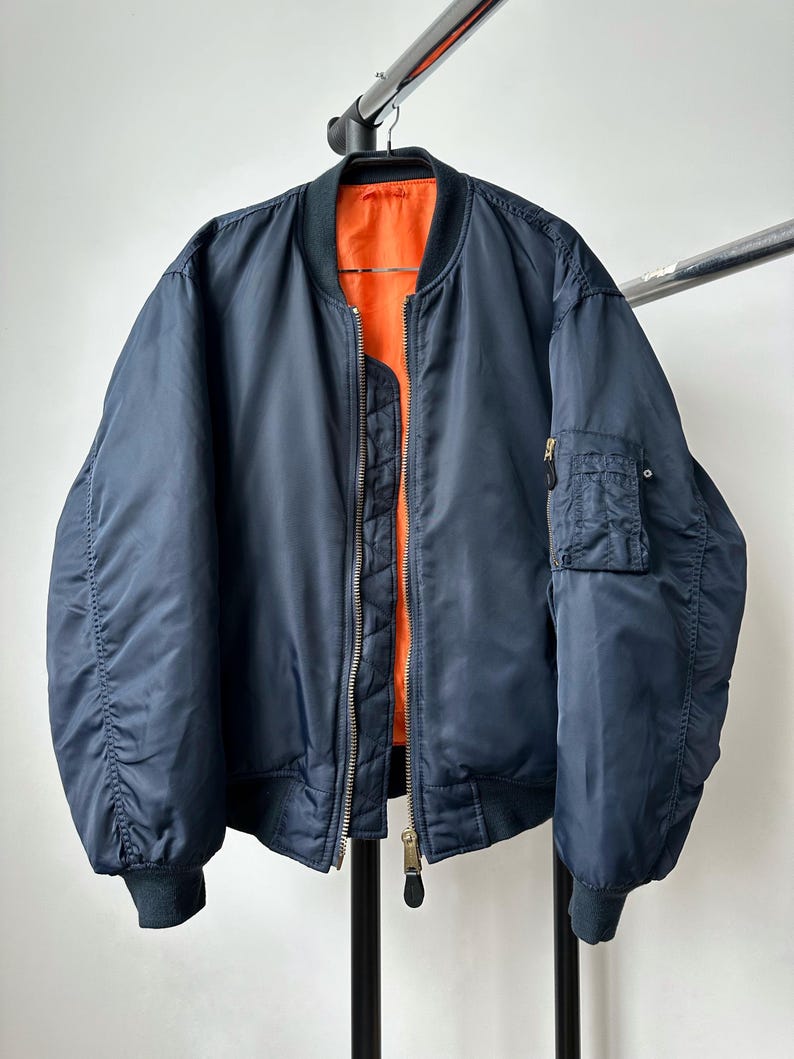 May include: Navy blue bomber jacket with a bright orange interior. The jacket features a zip-up front, a small pocket on the left sleeve, and ribbed cuffs and hem. The jacket is hanging on a black hanger.