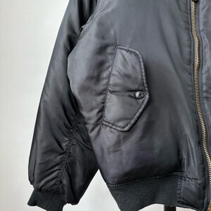 May include: A black bomber jacket featuring a smooth, satin-like finish, a gold-toned metal front zipper, and a snap-button flap pocket on the side. The jacket includes ribbed knit cuffs and a ribbed hem, with a bright orange lining visible at the collar.