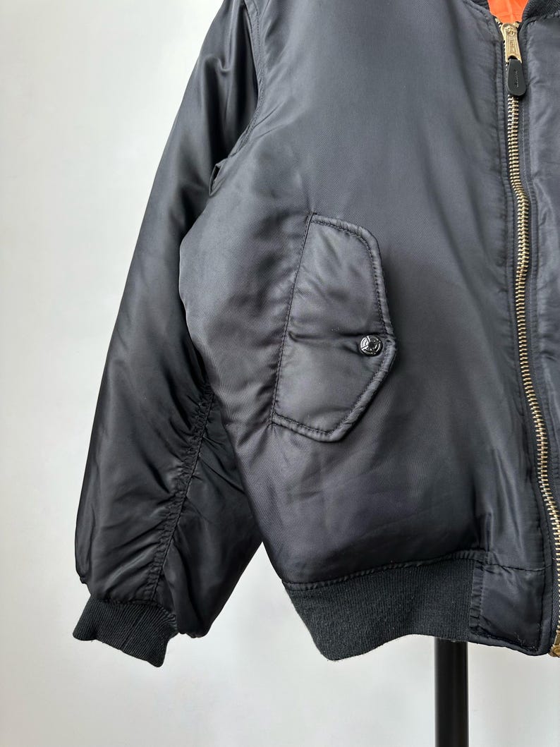 May include: A black bomber jacket featuring a smooth, satin-like finish, a gold-toned metal front zipper, and a snap-button flap pocket on the side. The jacket includes ribbed knit cuffs and a ribbed hem, with a bright orange lining visible at the collar.
