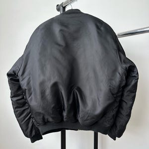 May include: A solid black bomber jacket featuring a ribbed collar, ribbed cuffs, and a ribbed hem. The jacket is constructed from a smooth, slightly reflective fabric and is displayed hanging on a metal hanger against a plain white background.