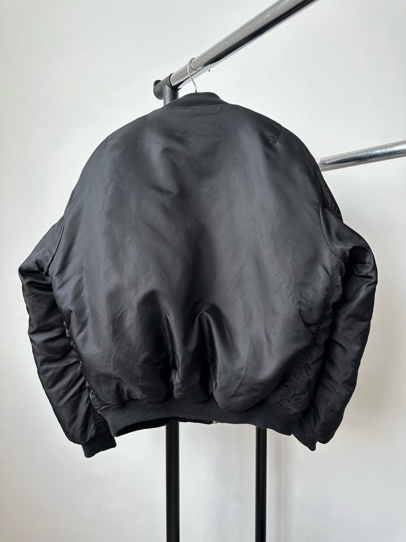 May include: A solid black bomber jacket featuring a ribbed collar, ribbed cuffs, and a ribbed hem. The jacket is constructed from a smooth, slightly reflective fabric and is displayed hanging on a metal hanger against a plain white background.