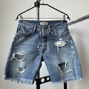 May include: Light blue denim shorts with distressed details, including rips and a frayed hem. The shorts are button-fly and feature the brand name "Acne" on the waistband. They are hanging on a black hanger against a neutral background.