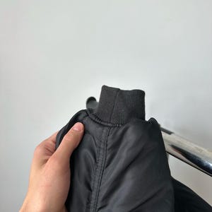 May include: A black bomber jacket sleeve featuring a ribbed knit cuff. The jacket is constructed from a smooth, slightly reflective synthetic fabric with visible vertical seam detailing running along the length of the sleeve toward the cuff.