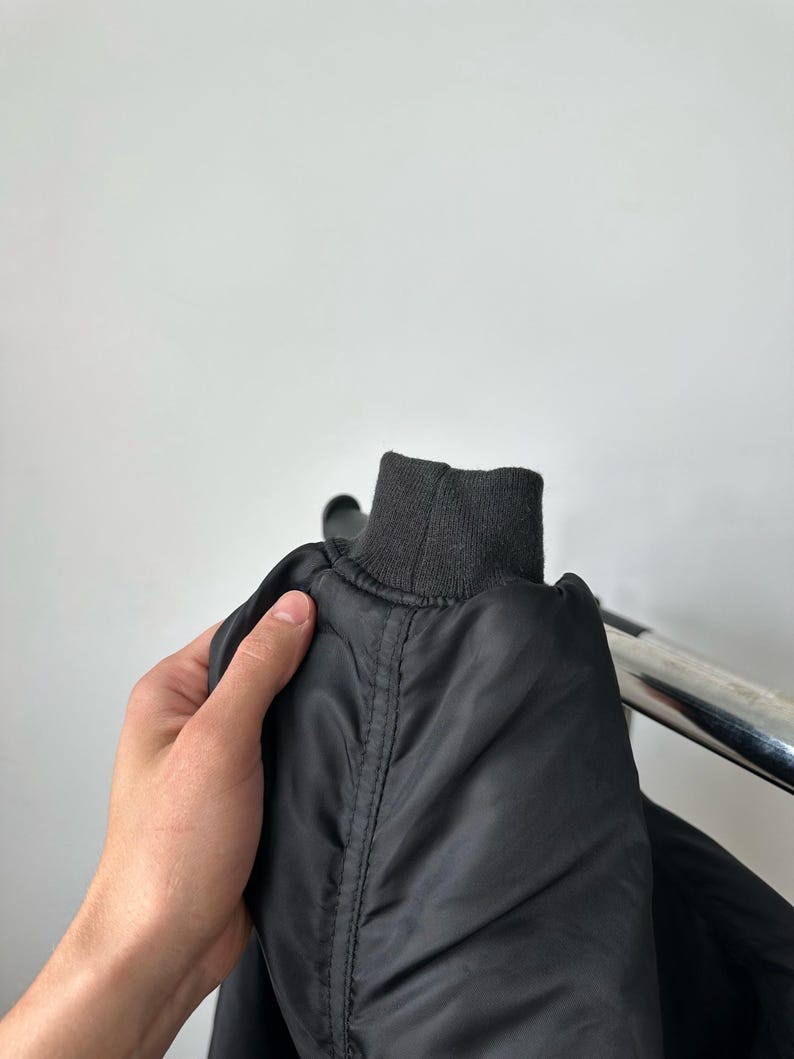 May include: A black bomber jacket sleeve featuring a ribbed knit cuff. The jacket is constructed from a smooth, slightly reflective synthetic fabric with visible vertical seam detailing running along the length of the sleeve toward the cuff.
