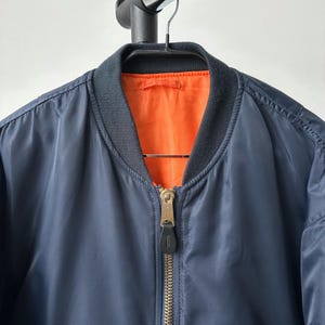 May include: Navy blue bomber jacket with a black ribbed collar and cuffs. The jacket has a gold zipper and an orange interior lining. The jacket is hanging on a black hanger.