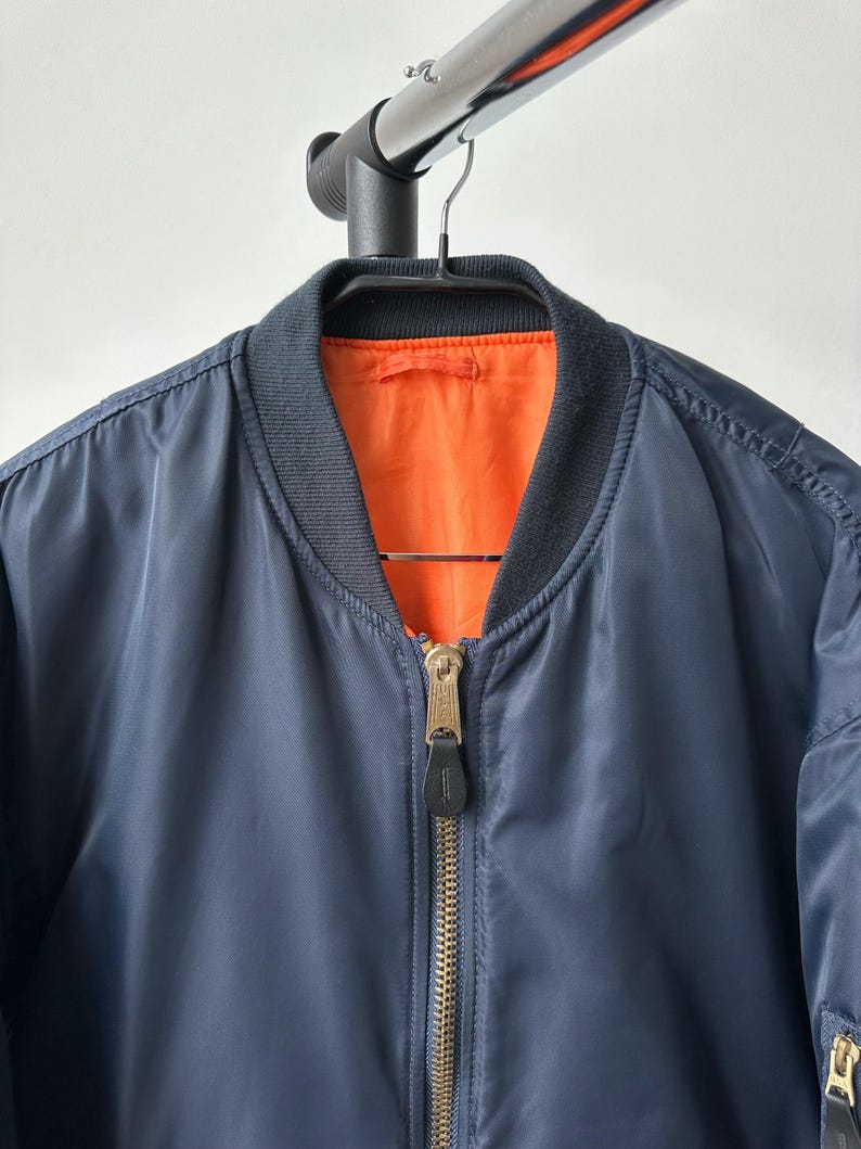 May include: Navy blue bomber jacket with a black ribbed collar and cuffs. The jacket has a gold zipper and an orange interior lining. The jacket is hanging on a black hanger.
