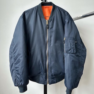 May include: A navy blue bomber jacket with a contrasting orange lining. The jacket features a ribbed collar, cuffs, and hem, a zip-up front, and a pocket on the left sleeve. The jacket is hanging on a metal rack.