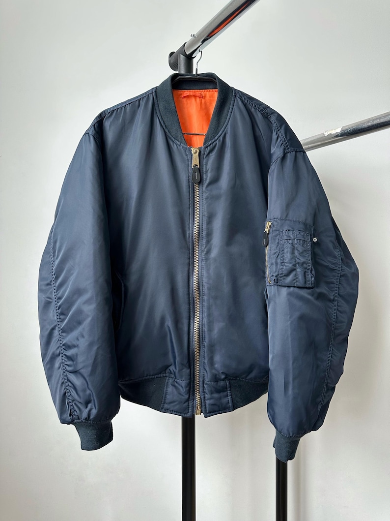 May include: A navy blue bomber jacket with a contrasting orange lining. The jacket features a ribbed collar, cuffs, and hem, a zip-up front, and a pocket on the left sleeve. The jacket is hanging on a metal rack.