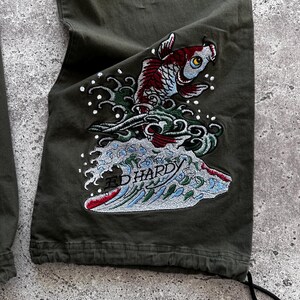 May include: Olive green cargo pants with a detailed embroidered design. The design features a koi fish in shades of red, blue, and white, emerging from stylized waves. The text "ED HARDY" is visible within the wave design.
