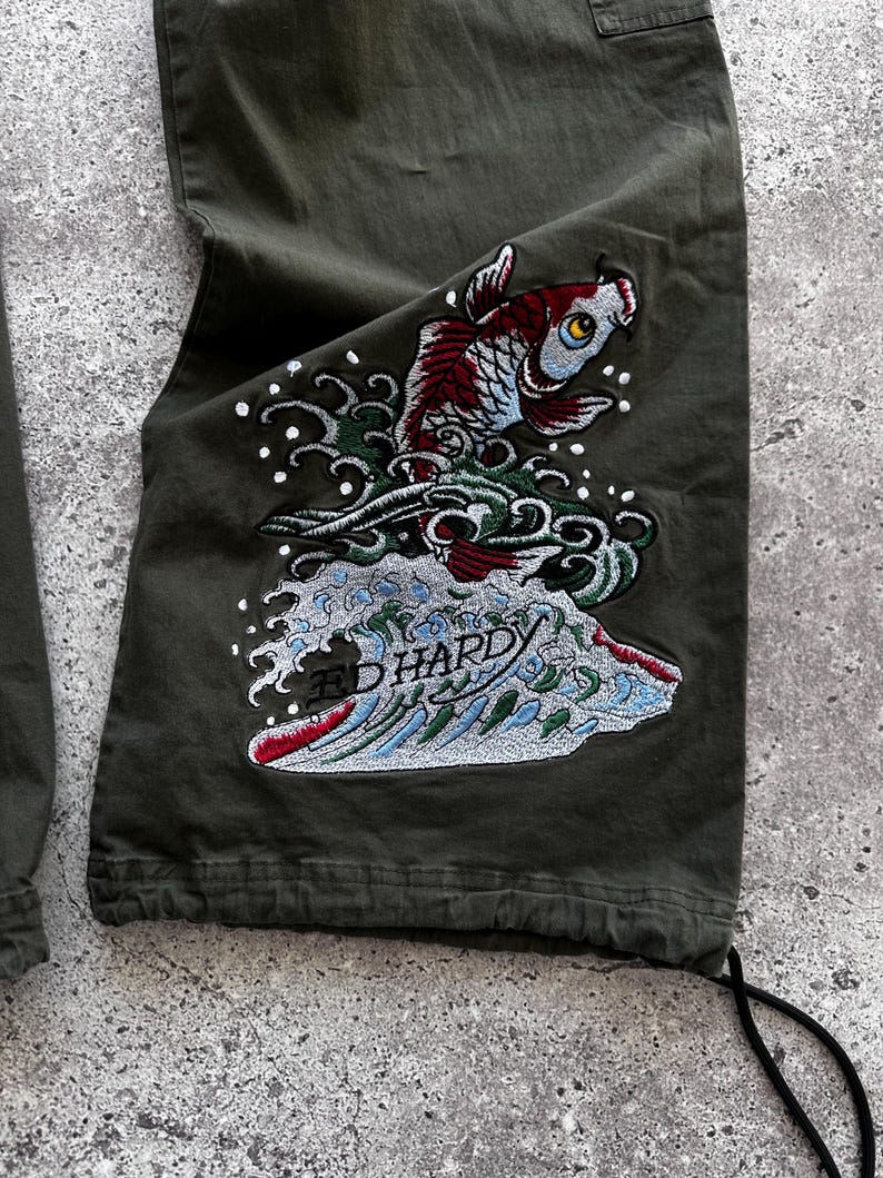 May include: Olive green cargo pants with a detailed embroidered design. The design features a koi fish in shades of red, blue, and white, emerging from stylized waves. The text "ED HARDY" is visible within the wave design.