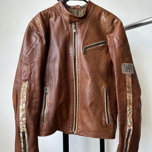 Puede incluir: Chaqueta de cuero marrón con cuello alzado, múltiples bolsillos con cremallera y parches decorativos. La chaqueta tiene una cremallera completa y rayas decorativas en las mangas. La chaqueta está colgada en una percha negra.