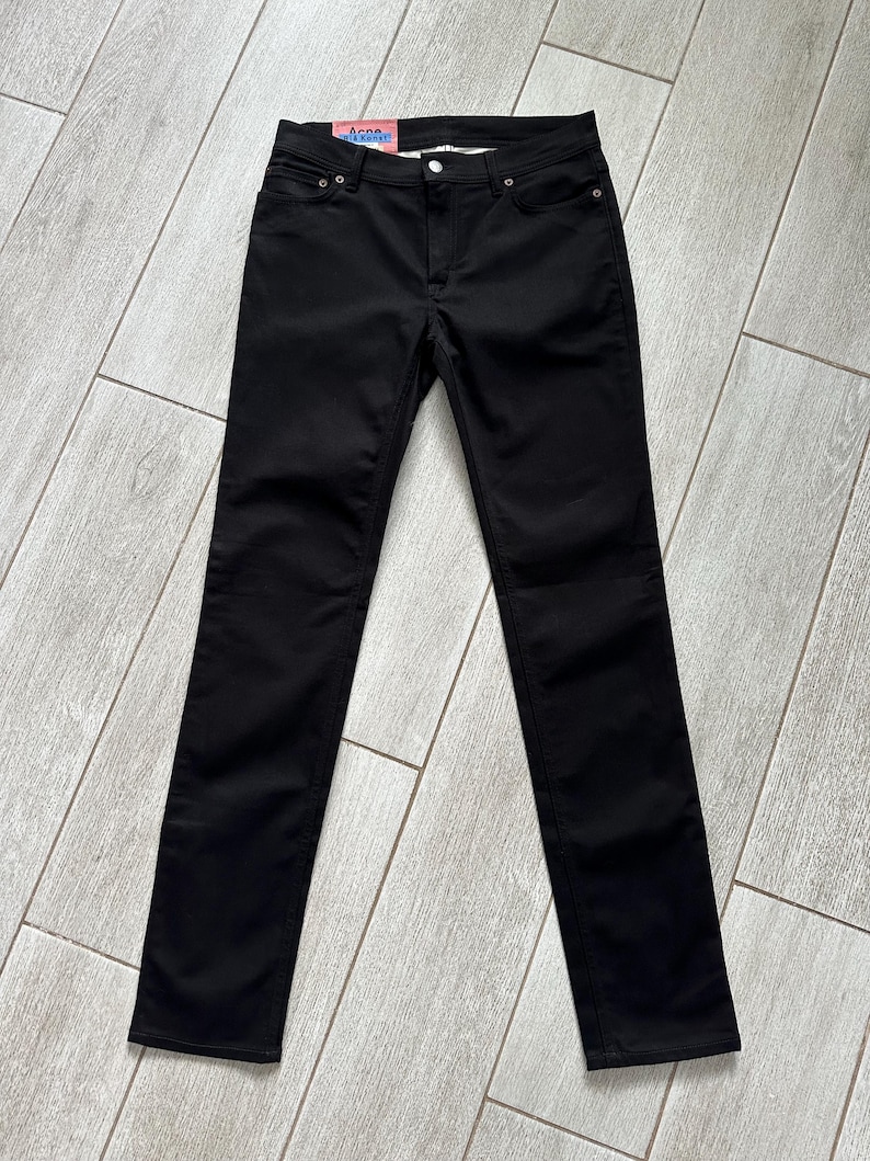 May include: A pair of solid black denim jeans featuring a classic five-pocket design with metal rivets and a button fly closure. The waistband includes a pink and blue brand label that reads Acne Studios, showcasing a slim-fit silhouette.