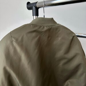 May include: Olive green bomber jacket hanging on a metal clothing rack. The jacket has a ribbed collar and a smooth, slightly shiny texture. The back of the jacket is visible, showing the seam details and the overall shape.