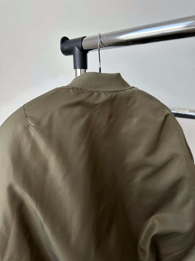 May include: Olive green bomber jacket hanging on a metal clothing rack. The jacket has a ribbed collar and a smooth, slightly shiny texture. The back of the jacket is visible, showing the seam details and the overall shape.