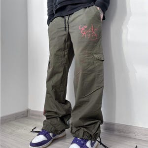 May include: Olive green cargo pants with the text "Ed 1979" in red script. The pants have multiple pockets and drawstring cuffs. The person is wearing white and purple sneakers with a brown sole.