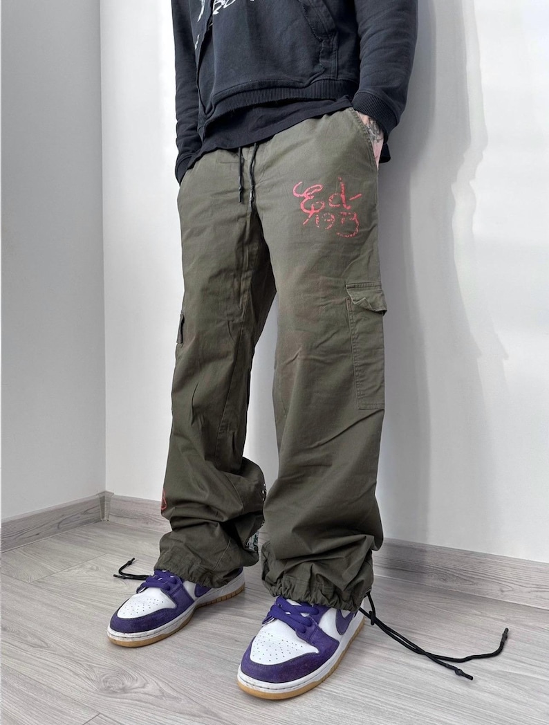 May include: Olive green cargo pants with the text "Ed 1979" in red script. The pants have multiple pockets and drawstring cuffs. The person is wearing white and purple sneakers with a brown sole.