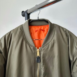 May include: Olive green bomber jacket with a bright orange lining. The jacket features a gold zipper and a black leather zipper pull. The jacket is hanging on a black hanger.