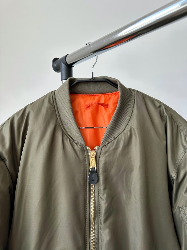 May include: Olive green bomber jacket with a bright orange lining. The jacket features a gold zipper and a black leather zipper pull. The jacket is hanging on a black hanger.