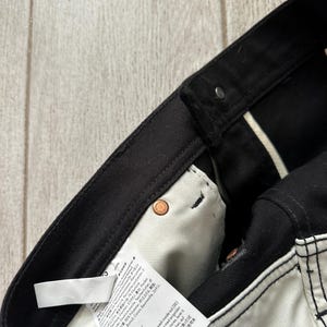 May include: Black denim pants featuring a white interior pocket lining with a care instruction label. The garment includes copper-colored metal rivets at the pocket corners and a black waistband with a button closure and belt loops.