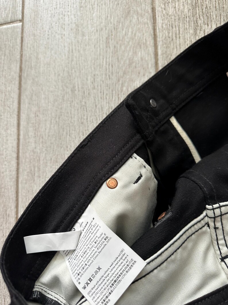 May include: Black denim pants featuring a white interior pocket lining with a care instruction label. The garment includes copper-colored metal rivets at the pocket corners and a black waistband with a button closure and belt loops.