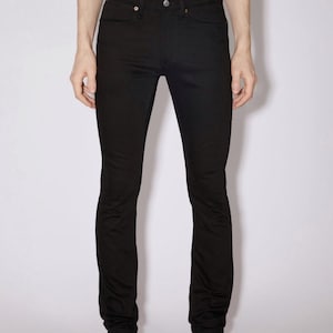 May include: Solid black denim jeans featuring a classic five-pocket design, a button fly closure, and a mid-rise waist. The text Acne Studios is printed in black at the top center of the image above the waistband of the black denim pants.