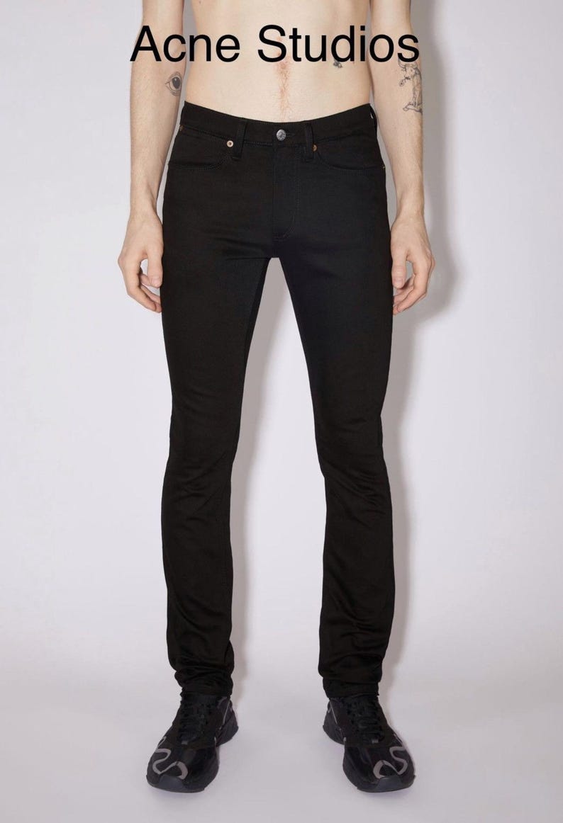May include: Solid black denim jeans featuring a classic five-pocket design, a button fly closure, and a mid-rise waist. The text Acne Studios is printed in black at the top center of the image above the waistband of the black denim pants.